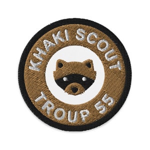 Op de afbeelding: Een bruin en zwart geborduurd patch met de tekst "Khaki Scout Troop 55" in wit. Een witte cirkel in het midden van de patch heeft een bruin wasbeergezicht.