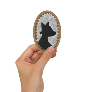 Puede incluir: Un parche ovalado marrón con un borde blanco bordado y una silueta negra de la cabeza de un perro en el interior.