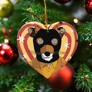 May include: Heart-shaped Christmas ornament featuring a cartoon animal with a black mask and spiral eyes. The ornament has a yellow and red striped background with white star accents. A gold string is attached for hanging.