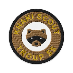 May include: A brown and black embroidered patch with the text "Khaki Scout Troop 55" in yellow. The patch features a cartoon raccoon in the center.