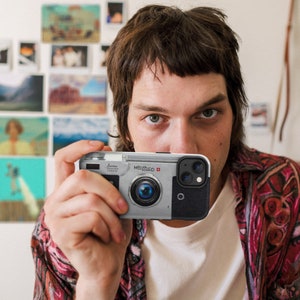 May include: A person holding a silver and black camera phone case with the text "MÖLLER-SCHMID" and a red Swiss flag. The case has a lens on the back that looks like an old camera lens.