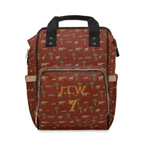 Puede incluir: Una mochila de lona marrón con estampado de animales de safari. La mochila tiene correas negras y cierre de cremallera. El texto "J.L.W. 7" está impreso en la parte delantera de la mochila.
