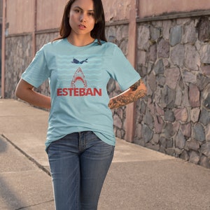 May include: A light blue t-shirt with a graphic of a shark's mouth with a diver inside. The text "ESTEBAN" is printed in red below the shark.