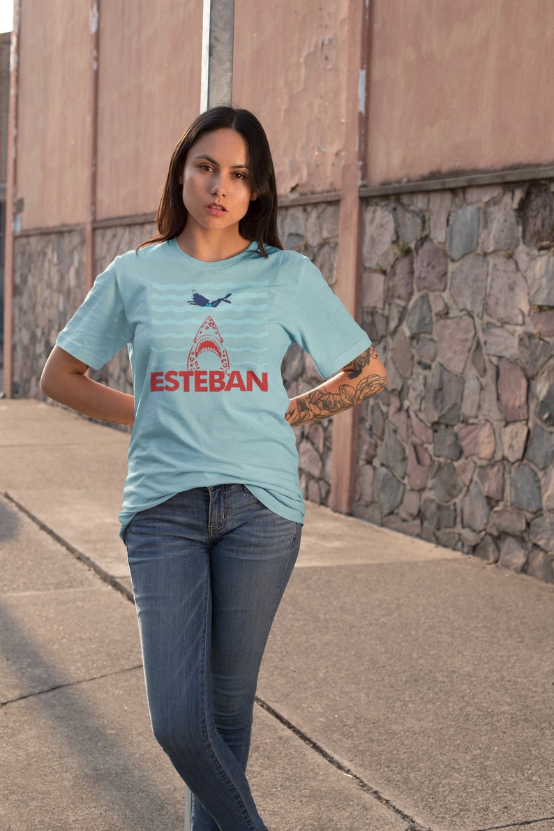 May include: A light blue t-shirt with a graphic of a shark's mouth with a diver inside. The text "ESTEBAN" is printed in red below the shark.