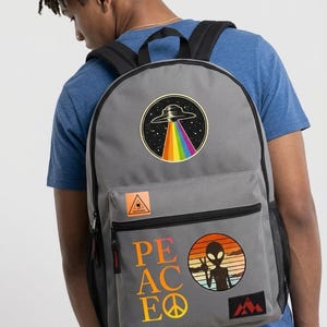 May include: A gray backpack featuring a circular patch of a UFO with a rainbow beam, a small orange square patch, the word PEACE with a peace sign for the letter O, and a circular patch of an alien making a peace sign against a sunset background.