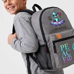 May include: A gray backpack featuring a graphic of a blue alien in a flying saucer holding a bouquet of flowers, with the text I COME IN PEACE above a front pocket. The word PEACE is printed in large, colorful letters on the lower front section.