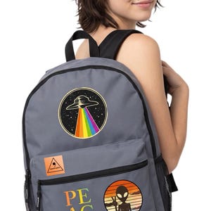 May include: A grey backpack featuring a circular patch of a UFO with a rainbow beam, a square orange patch with the text Apostrophe, the word PEACE with a peace sign, and a circular patch of an alien making a peace sign against a sunset.