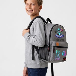 May include: A gray backpack featuring a graphic of an alien in a flying saucer holding flowers, with the text I COME IN PEACE. Below, the word PEACE is printed in a colorful gradient font, accompanied by a peace sign symbol and a small mountain icon.