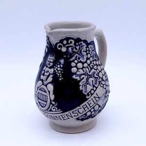 May include: A blue and white ceramic pitcher with a grapevine design and the word "SONNENSCHEIN" on the side. The pitcher has a handle and a round body.