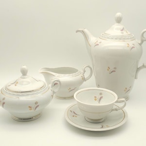 Może przedstawiać: Biały porcelanowy zestaw do herbaty z kwiatowym wzorem. Zestaw zawiera czajnik, dzbanek do śmietanki, cukiernicę, filiżankę i spodek.