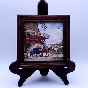 May include: A framed tile painting depicting a Dutch windmill scene with a horse-drawn cart and a man in the foreground. The tile is displayed on a wooden easel.