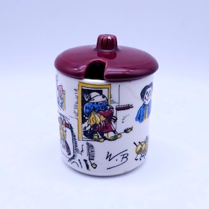 Puede incluir: Un tarro de cerámica blanco con tapa marrón y un diseño caprichoso de niños jugando. El tarro tiene un pequeño pico en la tapa y las iniciales "W.B." están escritas en el lateral.