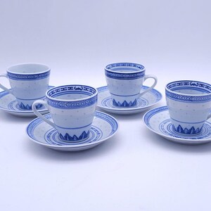 Op de afbeelding: Een set van vier witte keramische kopjes met bijpassende schoteltjes, elk versierd met een blauw patroon met een geometrische rand, kleine stippen over het lichaam en een gestileerd berglandschap aan de onderkant van de kopjes.