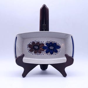 Op de afbeelding: Een rechthoekige keramische serveerschaal met een gespikkelde gebroken witte afwerking, voorzien van twee handgeschilderde bloemmotieven in bruin en blauw in het midden. De schaal heeft donkerblauwe accenten op de handgrepen en staat op een houten standaard.