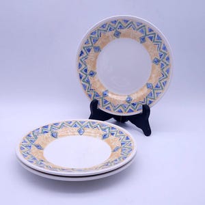 May include: Two white ceramic plates with a decorative border of blue, green, and tan geometric shapes. One plate is displayed on a black stand, while the other is stacked with a second plate.