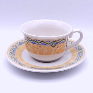 May include: A white ceramic teacup and saucer set. The cup and saucer have a decorative pattern of yellow, blue, and green geometric shapes. The cup has a curved handle and sits on a matching saucer.