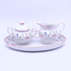 Könnte beinhalten: Ein weißes Porzellan-Zucker- und Milchkännchen-Set mit Blumendesigns, das auf einem weißen ovalen Tablett mit rosa Rand steht.