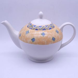 May include: White ceramic teapot with a lid and handle. The body of the teapot is white, with a tan band featuring blue and green geometric designs. The lid has a white knob.