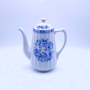 Op de afbeelding: Een witte theepot van keramiek met een blauw en gouden bloemenmotief. De theepot heeft een tuit en een handvat.