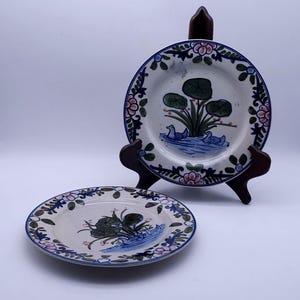 May include: Two round ceramic plates featuring a hand-painted design of two blue ducks swimming in water near green lily pads. The rim of each plate is decorated with a repeating pattern of pink flowers and dark green leaves on a white background.