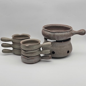 Puede incluir: Un juego de cinco ollas de fondue de cerámica marrón y gris. Las ollas están apiladas una encima de la otra, con la olla más grande en la parte inferior. Las ollas tienen un acabado moteado y están diseñadas para derretir queso o chocolate.