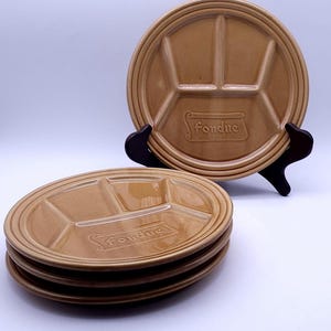 Op de afbeelding: Een set bruinbeige fondueborden. Elk bord heeft vier verdeelde secties en het woord "fondue" is in reliëf aangebracht. Eén bord staat op een zwarte standaard, de andere zijn opgestapeld.