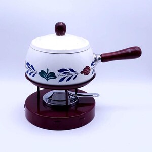 Op de afbeelding: Witte keramische fonduepot met een blauw en groen bloemenmotief en een houten handvat. De pot staat op een rode metalen standaard met een brander.