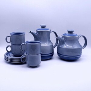 Pode incluir: Um conjunto de chá de cerâmica azul com uma chaleira, uma cafeteira, xícaras e pires. O conjunto tem um design de duas cores com uma faixa azul escura ao redor da base de cada peça. As xícaras estão empilhadas.