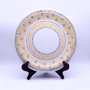 May include: A decorative ceramic plate with a cream-colored center and a border featuring a repeating pattern of blue diamonds and a yellow and blue design. The plate is displayed on a dark brown wooden stand.