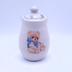 Op de afbeelding: Witte keramische pot met een blauw-wit deksel. De pot heeft een blauw-wit patroon en een bruine teddybeer met een blauwe strik.