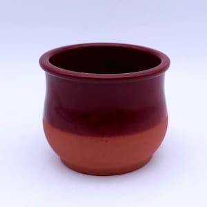 Puede incluir: Una pequeña maceta de cerámica hecha a mano con un esquema de color de dos tonos. La mitad superior es de un borgoña profundo, mientras que la mitad inferior es de color naranja terracota. La maceta tiene una forma redondeada con un borde ligeramente ensanchado.
