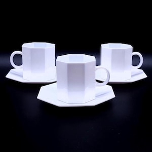 Peut inclure: Trois tasses à thé et soucoupes blanches de forme géométrique sont disposées sur une surface noire. Chaque tasse a une anse et repose sur une soucoupe assortie. Les tasses et les soucoupes ont un design moderne et minimaliste.