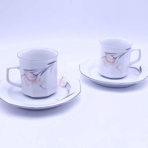 May include: Two white porcelain teacups with floral designs and matching saucers. The cups have a delicate floral pattern with pink and gray accents.