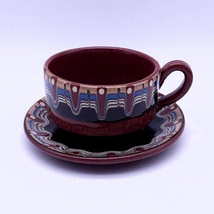 Puede incluir: Un juego de taza y platillo de cerámica. La taza y el platillo son de color burdeos oscuro con un patrón decorativo de líneas onduladas azules, doradas y blancas. La taza tiene un asa redondeada.