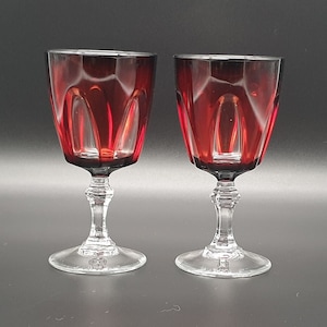 Può includere: Due bicchieri da vino in vetro trasparente con una tonalità rosso intenso. I bicchieri hanno una ciotola larga e uno stelo alto e sottile.