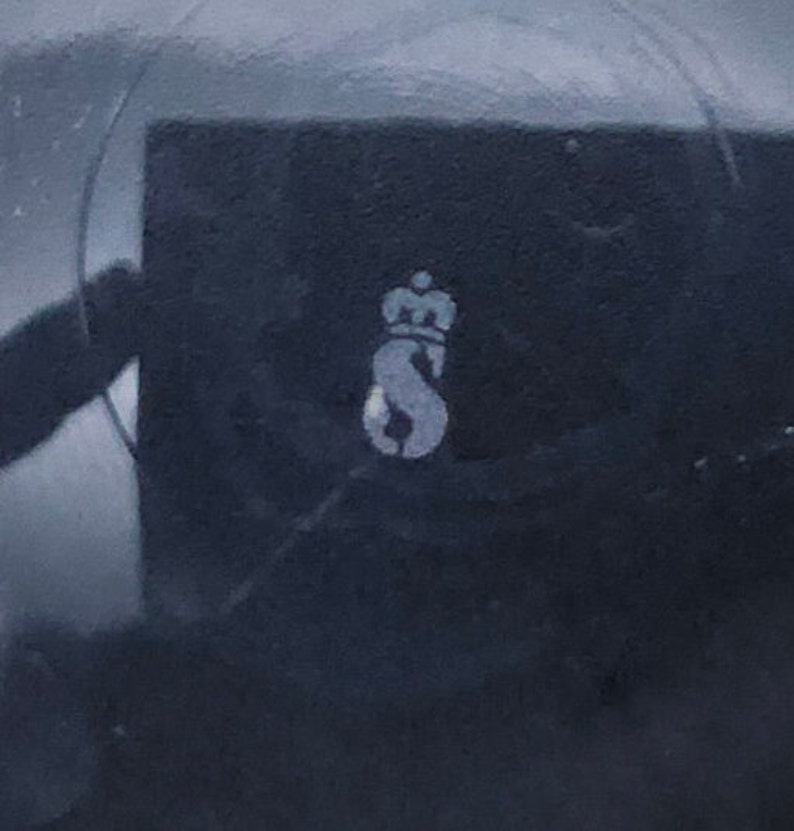 Peut inclure: Gros plan sur une surface noire avec une lettre 'S' stylis&eacute;e blanche surmont&eacute;e d'une couronne.