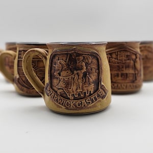 May include: A set of ceramic mugs with embossed designs. The front mug features a shield with a knight on horseback and the text "WARWICK CASTLE". The mugs are tan with brown accents and handles.