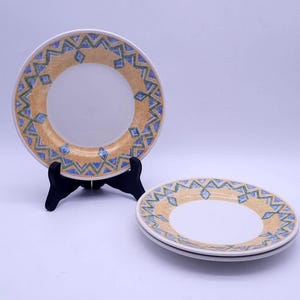 May include: Three white ceramic plates with a decorative border. The border features a repeating pattern of blue and green geometric shapes on a tan background. One plate is displayed on a black plate stand.