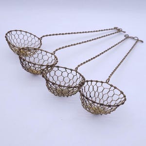 Op de afbeelding: Vier kleine, ronde, komvormige zeven gemaakt van dun, donker metalen gaas. Elke zeef heeft een zeshoekig gaaspatroon en is bevestigd aan een lange, slanke, gedraaide draadsteel die eindigt in een kleine ronde lus om op te hangen.