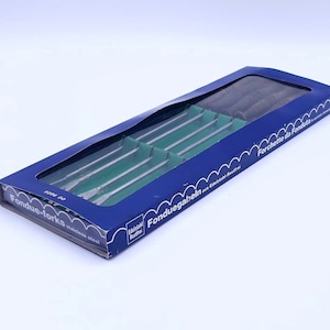 Può includere: Una scatola blu di forchette da fonduta in acciaio inossidabile. La scatola ha una finestra trasparente che mostra le forchette e divisori verdi. Il testo sulla scatola recita "Fondue-forks stainless steel" e "Forchette da Fonduta".