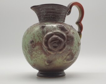 Könnte beinhalten: Eine grüne und braune Keramikkanne mit einem erhabenen Blumendesign und einem roten Henkel.