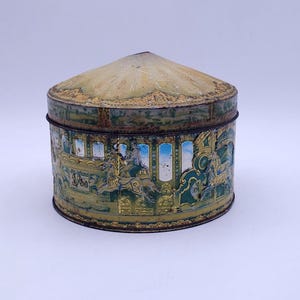 Op de afbeelding: Vintage ronde blikken doos met een conisch deksel. De blikken doos is versierd met een carrouselscène in tinten groen, goud en blauw. Het deksel heeft een verbleekte geelgroene kleur. De blikken doos lijkt verouderd.