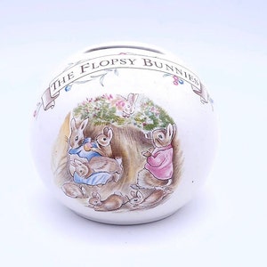 以下が含まれることがあります： ビートリックス・ポターの絵本「フロプシーの仲間たち」のカラフルなイラストが描かれた白いセラミック製の貯金箱。貯金箱には「The Flopsy Bunnies」という文字が書かれたバナーが付いています。