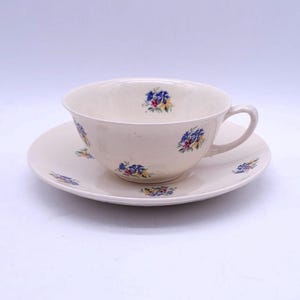 May include: A cream-coloured teacup and saucer set. The cup and saucer are decorated with small blue, yellow, and red floral patterns. The cup has a curved handle and sits on a matching saucer.