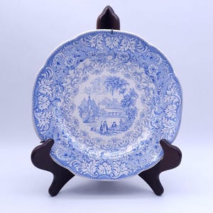 Op de afbeelding: Een ronde keramische bord met een blauw-wit transferpatroon. Het midden toont een landschap met figuren, gebouwen en een brug, terwijl de brede rand is voorzien van ingewikkelde bloemmotieven en blauwe bladeren in krulvorm.