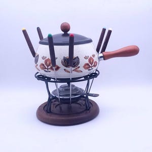 Op de afbeelding: Een witte keramische fonduepot met een bruin bloemenontwerp en een houten handvat. De pot staat op een zwarte metalen standaard met een houten basis en bevat acht fonduevorken met gekleurde handvatten.