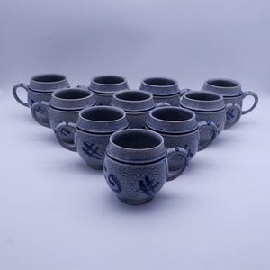 Może przedstawiać: Zestaw dziesięciu jasnoszarych ceramicznych kubków ułożonych w trójkąt. Każdy kubek ma teksturowaną, nakrapianą powierzchnię, gładki brzeg, zakrzywione ucho oraz ciemnoniebieskie, ręcznie malowane abstrakcyjne wzory na zaokrąglonym korpusie.