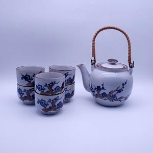 May include: A white ceramic teapot with a brown lid and a woven handle, along with four matching teacups with a blue and brown floral design.