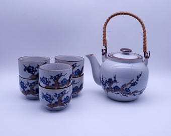 May include: A white ceramic teapot with a brown lid and a woven handle, along with four matching teacups with a blue and brown floral design.