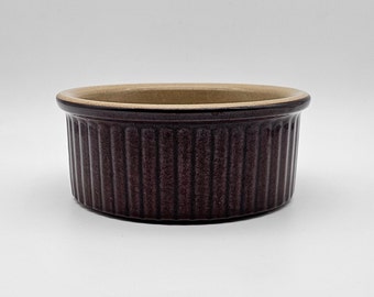 May include: A small, round, brown ceramic ramekin with a light brown interior. The ramekin has vertical ridges around the exterior.